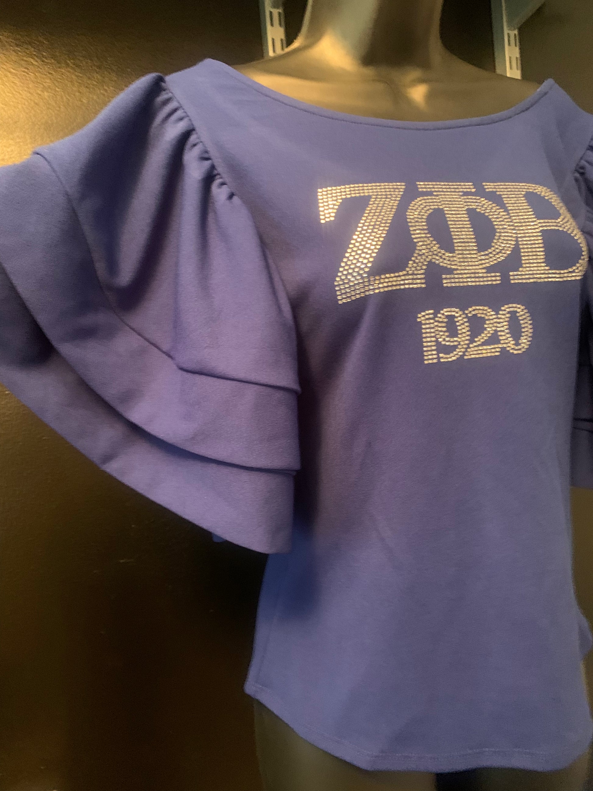 Shine with Zeta pride in the Zeta Bling Royal Blue Blouse. These vibrant royal blue top features elegant, ruffled sleeves and sparkling bling details that make it perfect for Soror events or stylish outings.